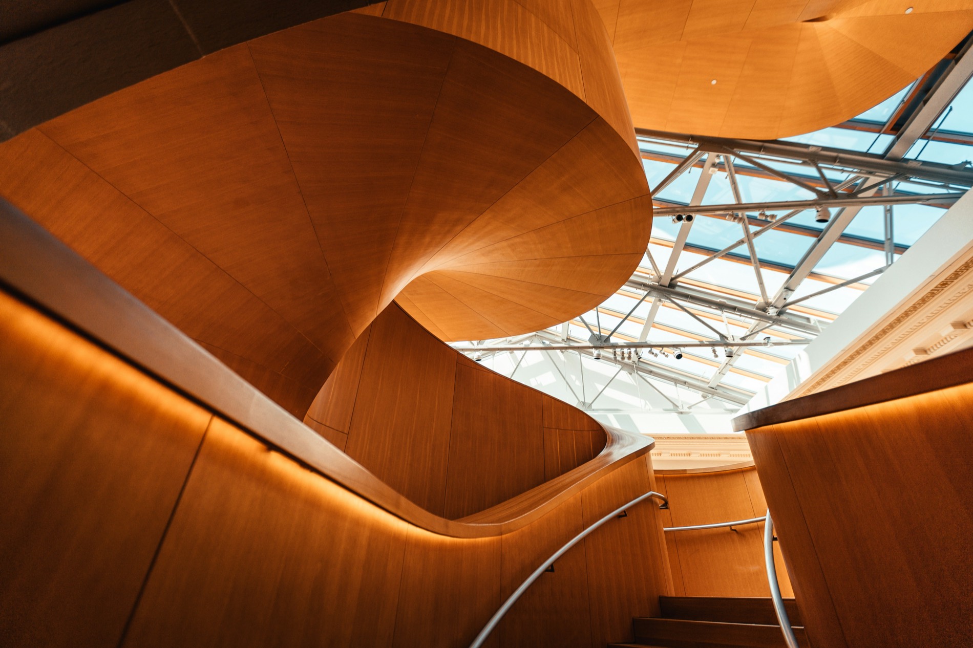 Sunlit curved wooden staircase beneath a glass atrium roof