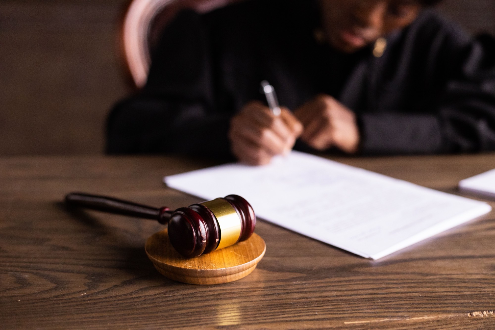 A wooden gavel on a desk as a judge signs a document in the background